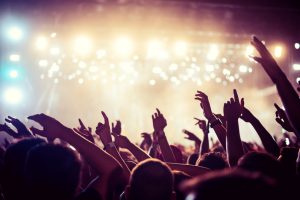 Audience with hands raised at a music festival and lights streaming down from above the stage. Soft focus, high ISO, grainy image.